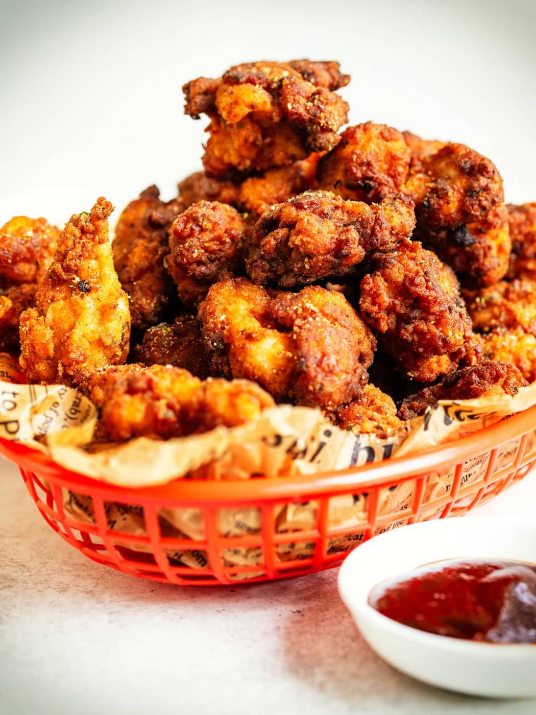 A red basket lined with paper is filled with crispy fried popcorn chicken, KFC-style. In the foreground, there is a small dish of red dipping sauce. The background is softly blurred.