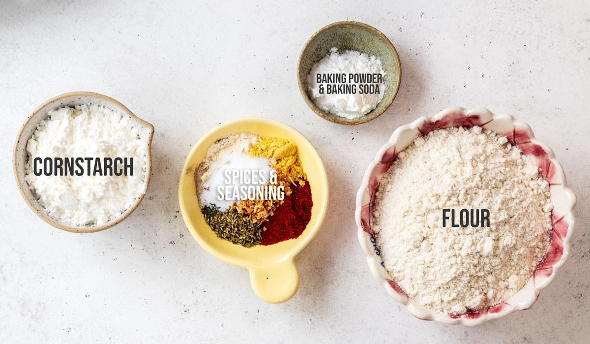 Four bowls containing labeled ingredients: cornstarch, flour, baking powder & baking soda, and a mix of colorful spices and seasoning, arranged on a light surface.