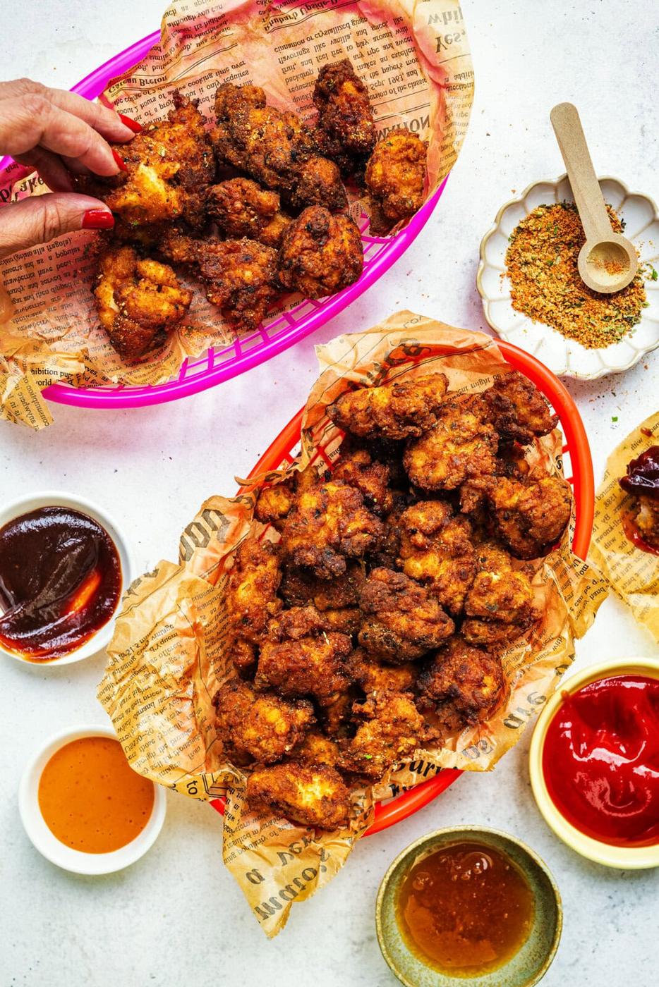 Two baskets lined with newspaper-style paper hold crispy fried cauliflower bites. Sauces and seasoning are in small bowls nearby, and a hand reaches for a piece from one basket.