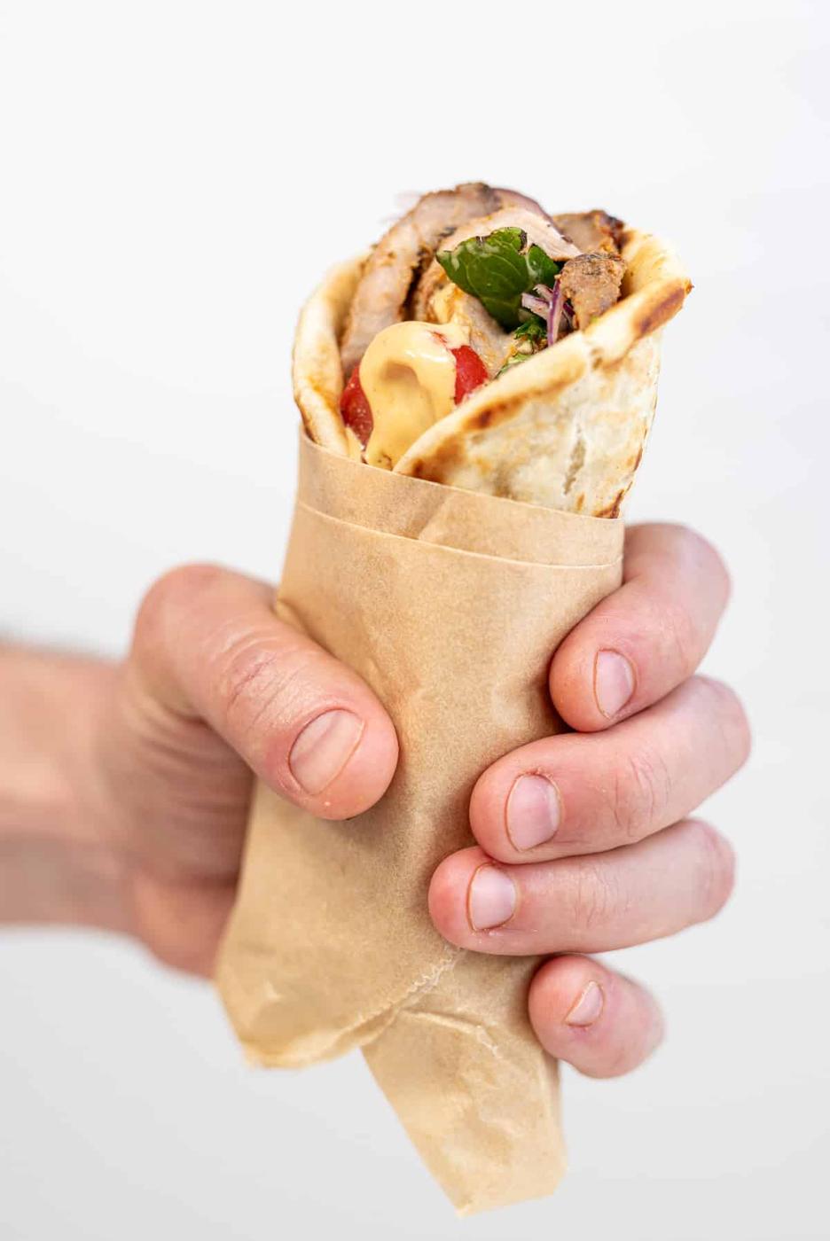 A hand holds a wrapped pita filled with air fryer pork gyros, leafy greens, sliced tomato, and sauce—an irresistible twist on an authentic Greek recipe—against a plain white background.