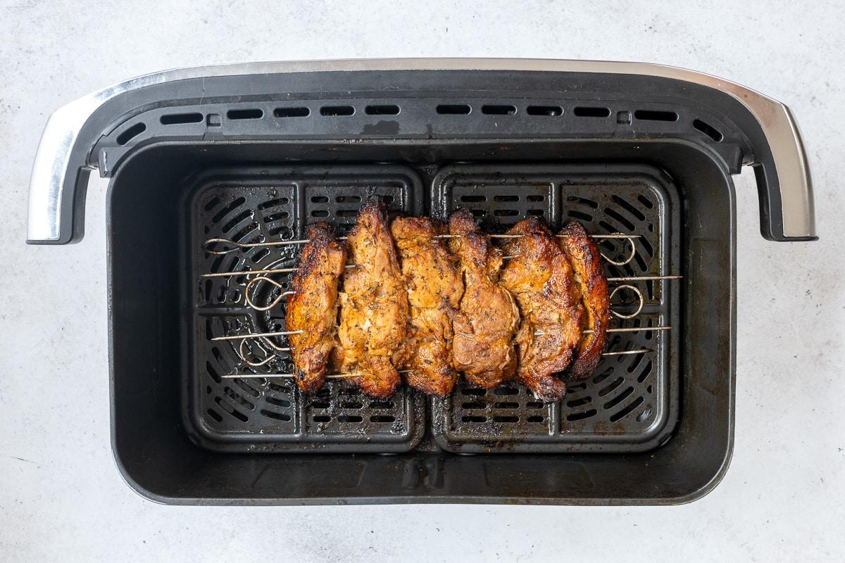 Five skewers with seasoned, grilled meat for Air Fryer Pork Gyros are placed inside an air fryer basket on a light surface, showing a cooked, golden-brown exterior reminiscent of an authentic Greek recipe.