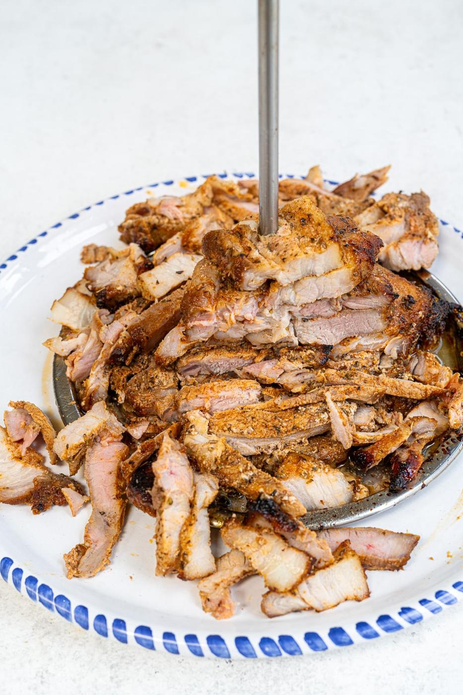 A large plate of sliced, seasoned Air Fryer Pork Gyros stacked on a vertical skewer, ready to serve in true authentic Greek recipe style, with a blue and white rimmed plate on a light surface.