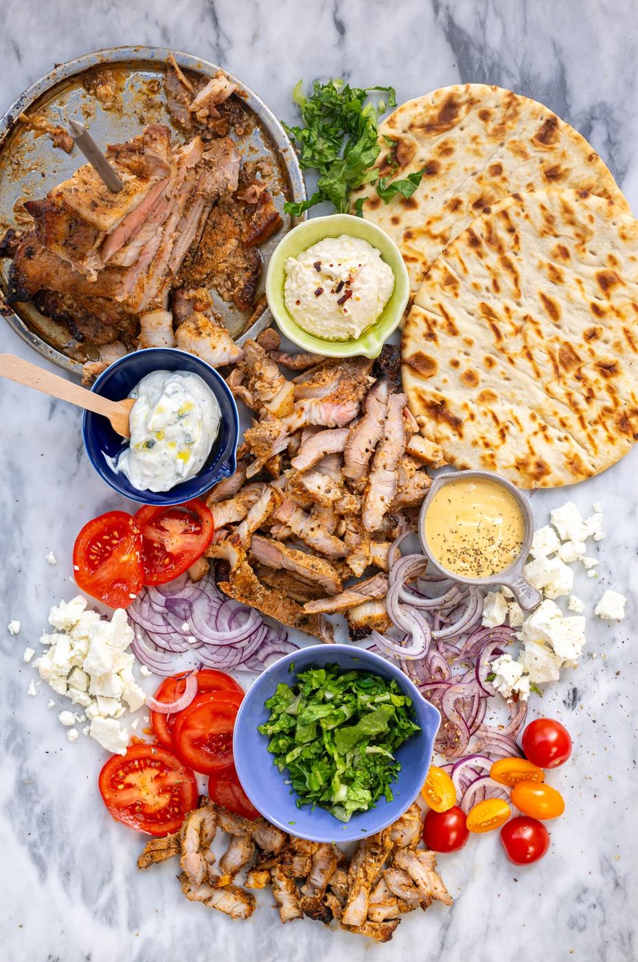 Deconstructed Air Fryer Pork Gyros ingredients on a marble surface: grilled meat, pita bread, tzatziki, hummus, mustard sauce, sliced tomatoes, red onions, feta cheese, leafy greens, chopped herbs, and cherry tomatoes.