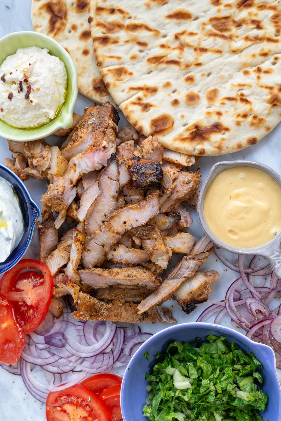 A platter featuring Pork Gyros with sliced grilled meat, two pieces of flatbread, sliced red onions, tomato, chopped lettuce, a bowl of yellow sauce, tzatziki, and a bowl of whipped feta cheese—an authentic Greek recipe delight.