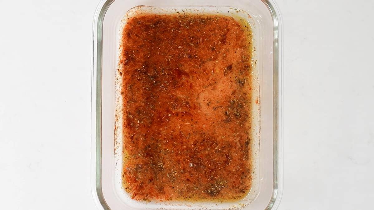 A glass baking dish containing a reddish-brown marinade with visible herbs and spices, perfect for preparing authentic Greek recipe Pork Gyros, viewed from above on a white background.