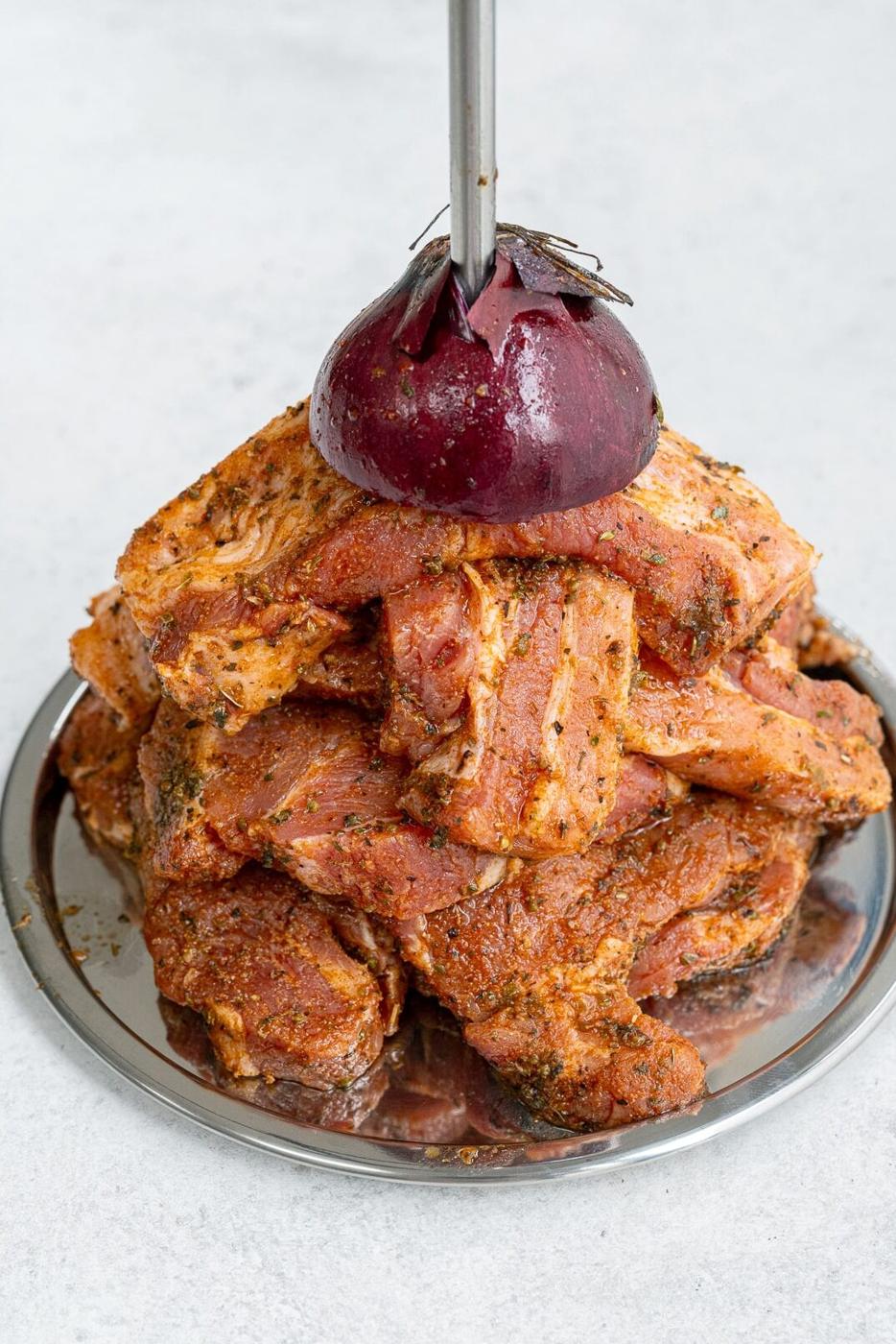 A stack of marinated pork gyros slices arranged on a round metal tray, topped with half a red onion skewered by a vertical metal rod, captures the essence of an authentic Greek recipe.