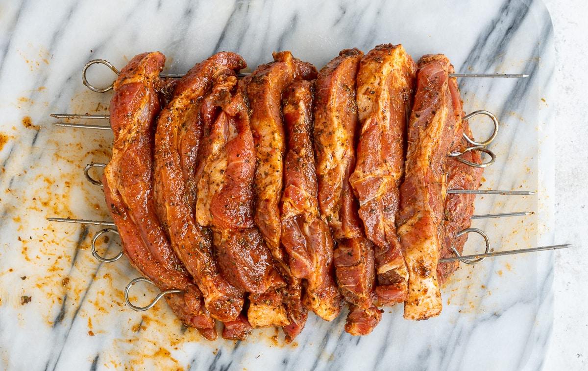 Raw marinated pork skewers, prepared for grilling, are arranged side by side on a marble surface. Seasoned with spices and placed on metal skewers, these are perfect for making authentic Greek recipe Pork Gyros.