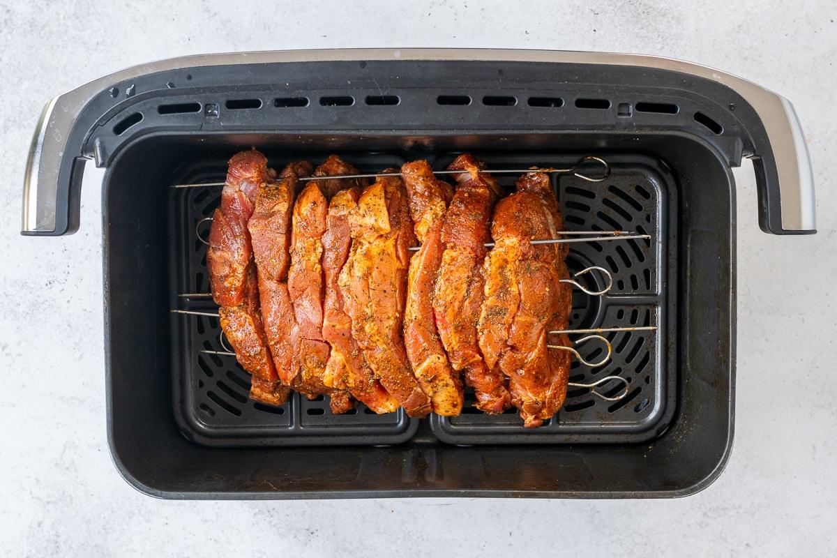 Raw, seasoned meat slices for authentic Greek recipe Pork Gyros are skewered on metal sticks and arranged in an air fryer basket, ready for cooking. The spiced meat promises delicious Air Fryer Pork Gyros, set on a light-colored surface.
