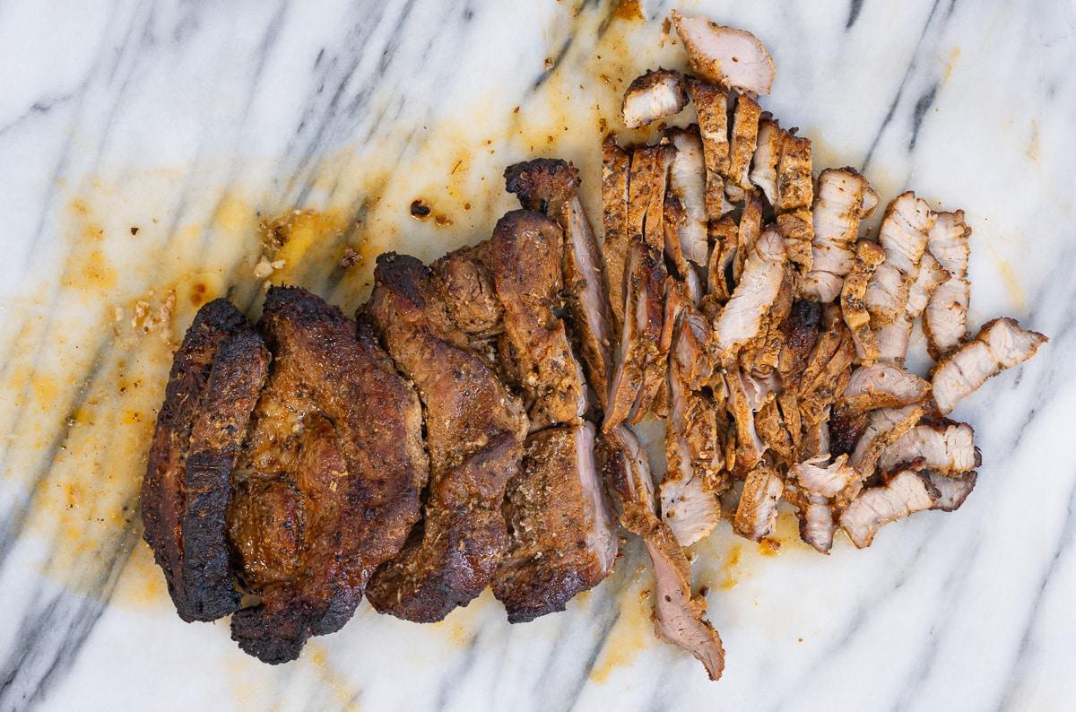 A cooked, seasoned pork roast rests on a white marble surface, with part of it sliced into thin strips—perfect for Pork Gyros. The meat looks juicy, with visible char and spices inspired by an authentic Greek recipe.