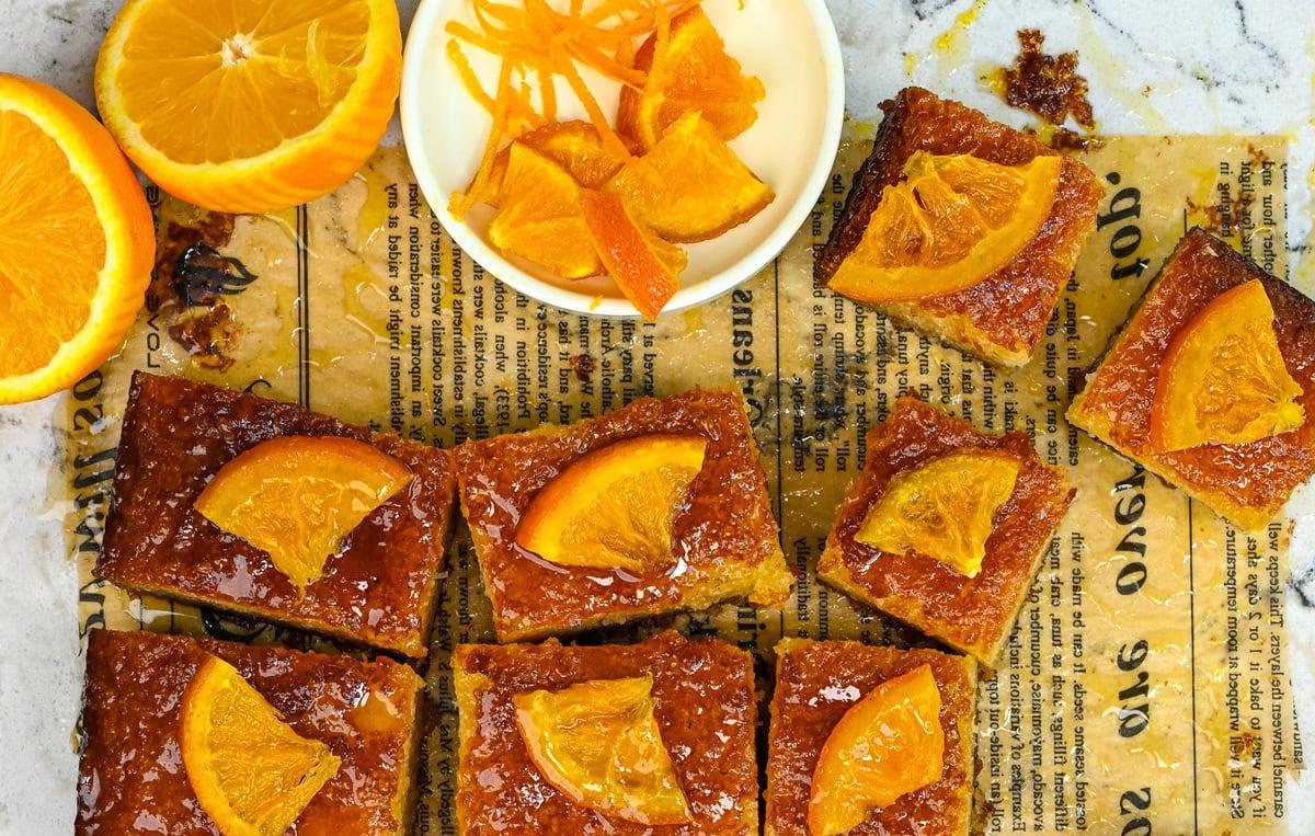 Nine pieces of Greek orange cake topped with orange slices, arranged on parchment paper with text, next to a sliced orange and a small dish holding more orange slices and zest.