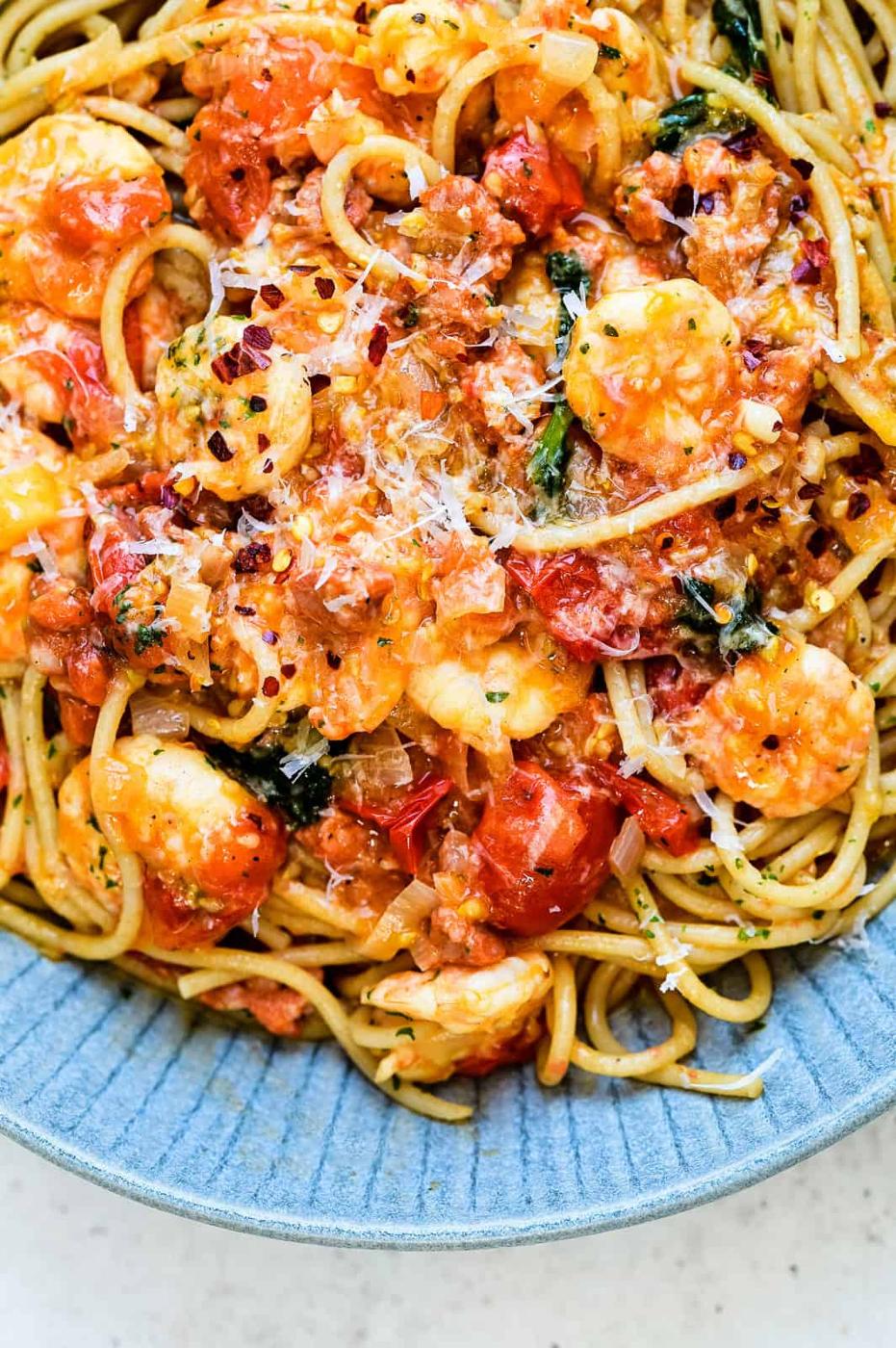 A close-up of prawn pasta topped with shrimp, chopped tomatoes, spinach, grated cheese, and red pepper flakes, served in a blue bowl.