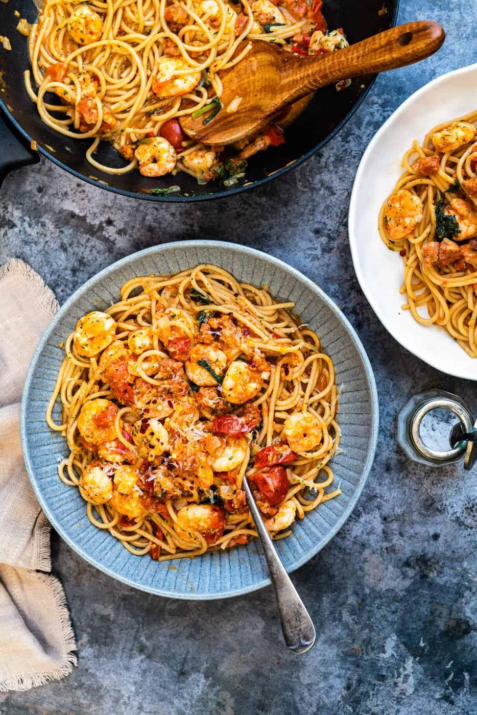 A bowl of delicious prawn pasta sits next to a skillet and another plate of pasta. A fork rests in the bowl, and a glass jar is nearby on a textured gray surface—perfect inspiration for your next prawn pasta recipe.