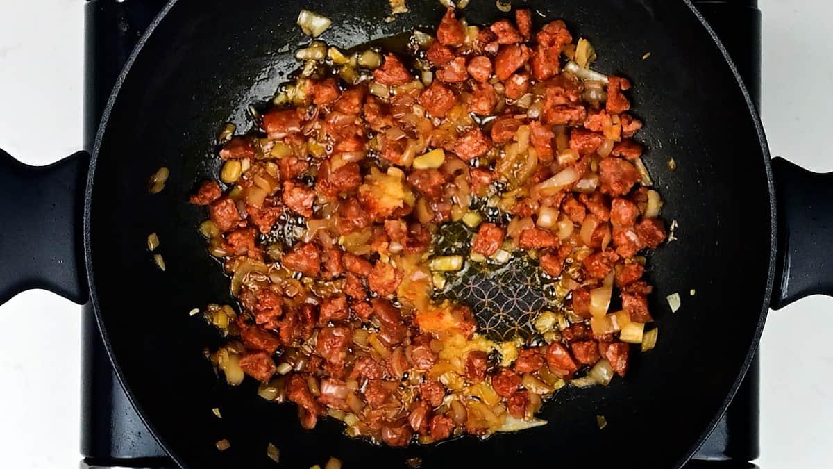A skillet with diced chorizo, chopped onions, and garlic sautéing together for a delicious prawn and chorizo pasta. The mixture of browned meat and softened vegetables is the perfect base for your next prawn pasta recipe. The pan sits on a stovetop.
