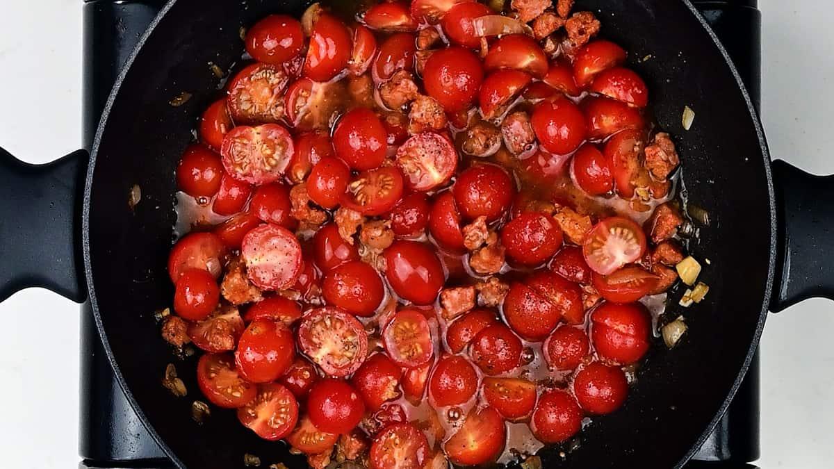 A black pan filled with halved cherry tomatoes and crumbled sausage cooks together in sauce, reminiscent of the vibrant flavors found in a classic prawn and chorizo pasta. Seen from above.