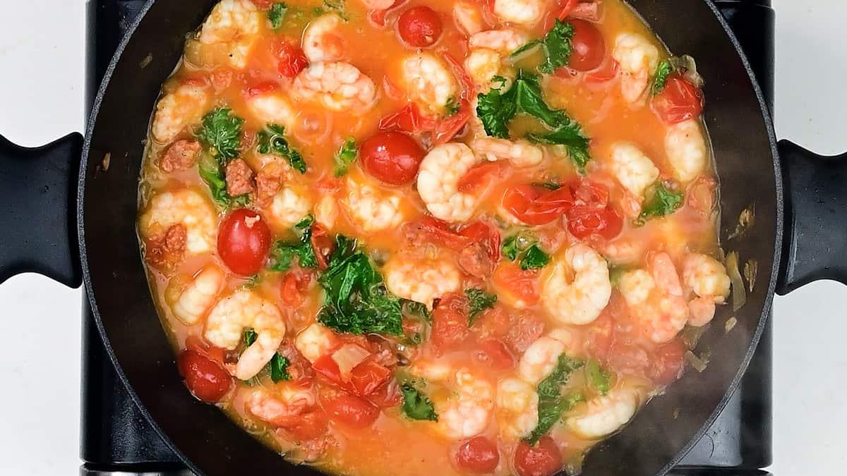 A skillet filled with shrimp, cherry tomatoes, chopped greens, and a light tomato-based sauce simmers on the stovetop—perfect for a quick prawn pasta recipe.
