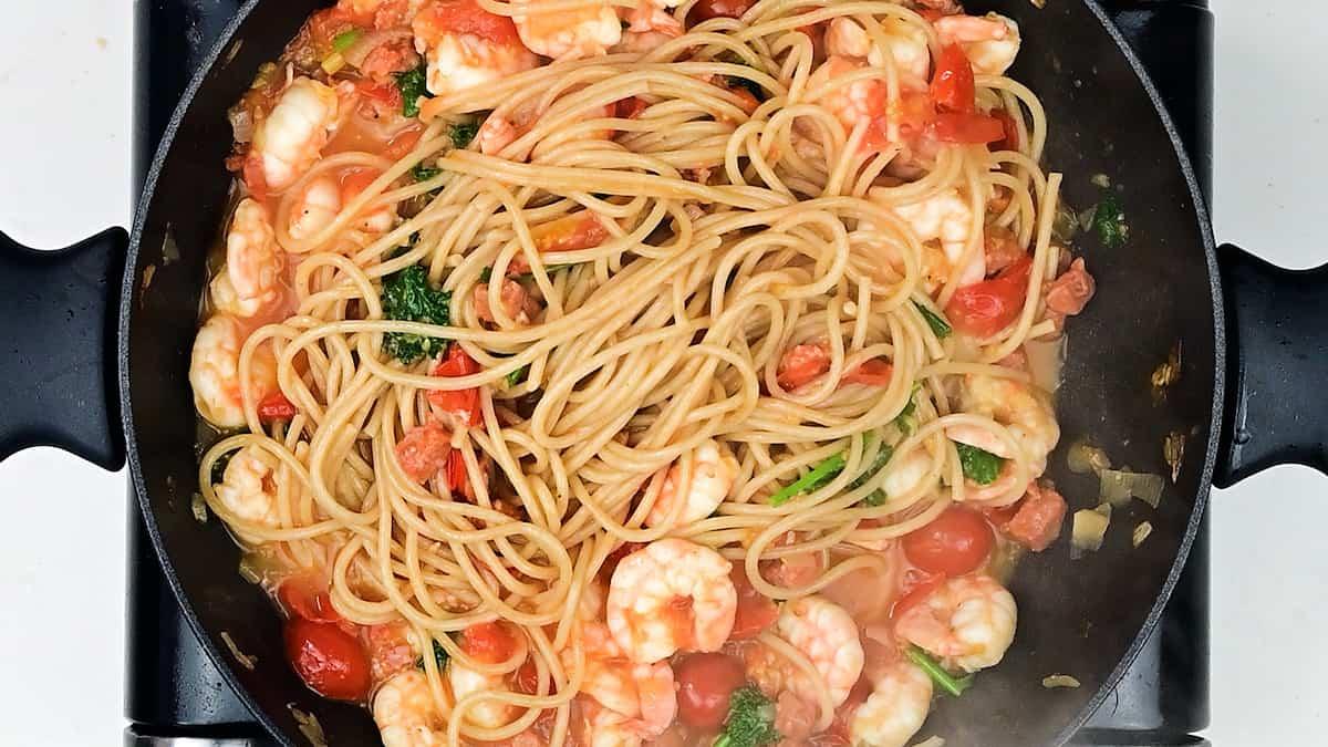 A pan of Prawn Pasta mixed with shrimp, chopped tomatoes, and green herbs sits on the stovetop, with steam rising from the hot dish.