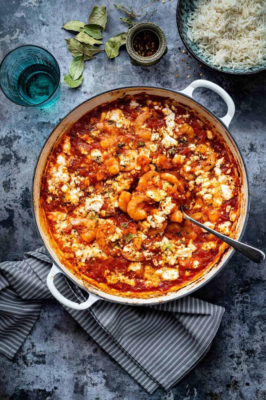A white pot filled with shrimp saganaki in a rich tomato sauce, garnished with herbs and crumbled cheese. A spoon rests inside the pot. Nearby are a bowl of rice, a glass of water, herbs, and a striped kitchen towel.