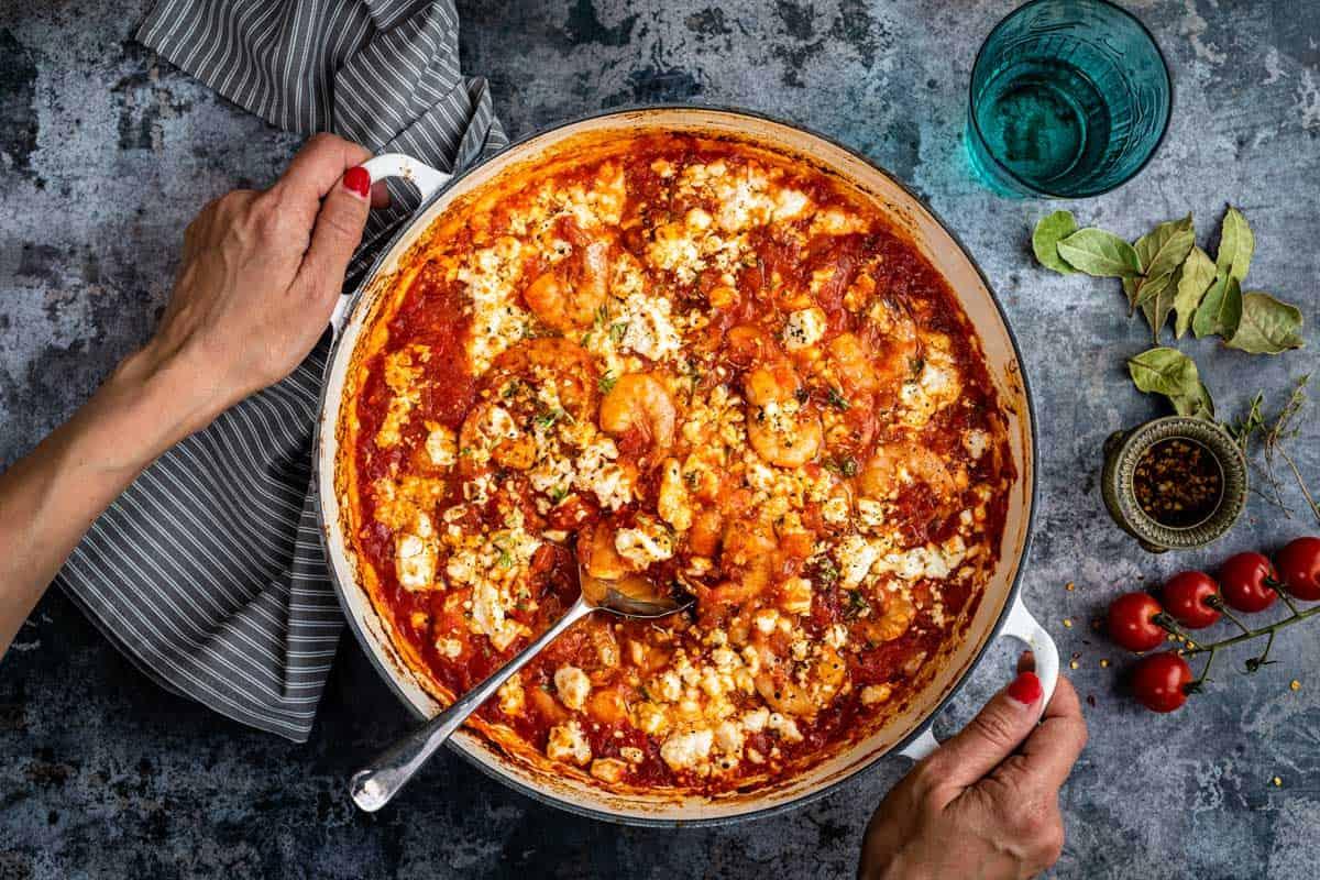 A person holds a pan of Prawn Saganaki—shrimp in tomato sauce topped with crumbled feta—set on a rustic surface with a striped towel, fresh herbs, cherry tomatoes, and a glass of water nearby.