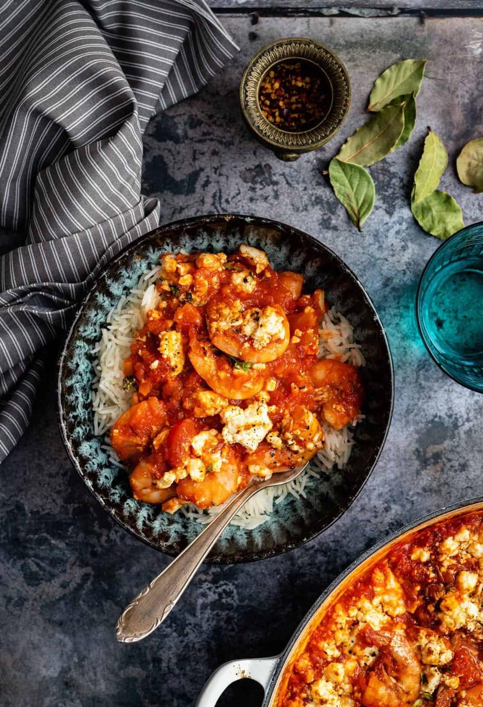 A bowl of rice topped with shrimp Saganaki—shrimp in a tomato-based sauce and crumbled feta—with a fork. Bay leaves, a small bowl of spices, and a blue glass rest nearby on a textured surface with a striped napkin.