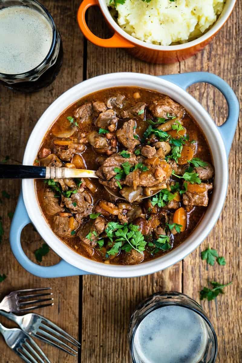 A bowl of hearty Irish beef stew garnished with fresh parsley, surrounded by mashed potato, two glasses of dark beer, and two forks on a rustic wooden table.