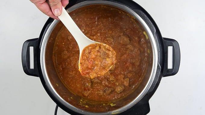 A hand holds a white ladle filled with thick, chunky Lamb Dhansak stew over an open Instant Pot containing more of the flavorful Lamb Dhansak recipe.