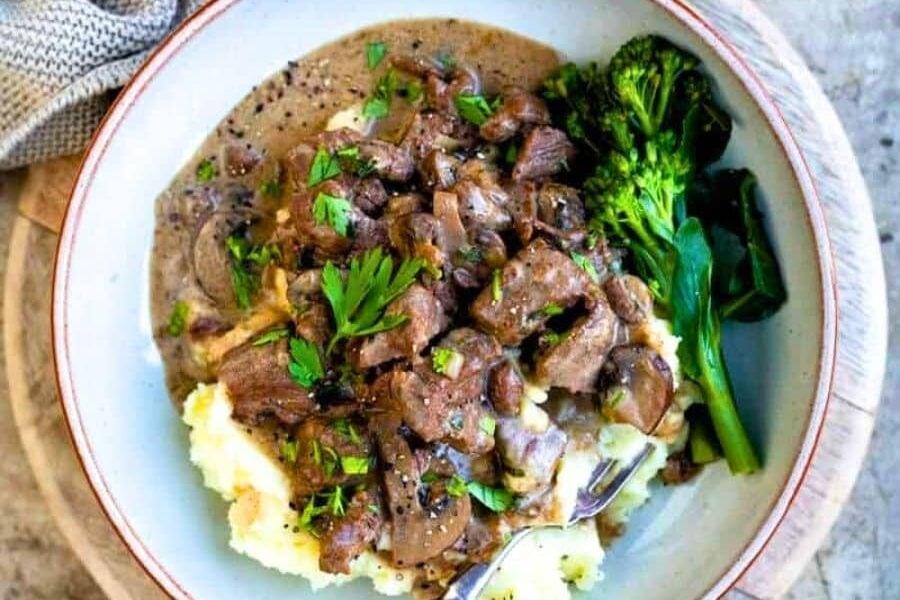 A bowl of creamy steak Diane with mushrooms and parsley served over mashed potatoes, alongside steamed broccoli. A hand holds a fork in the bowl. Salt and pepper shakers and a cloth are in the background.
