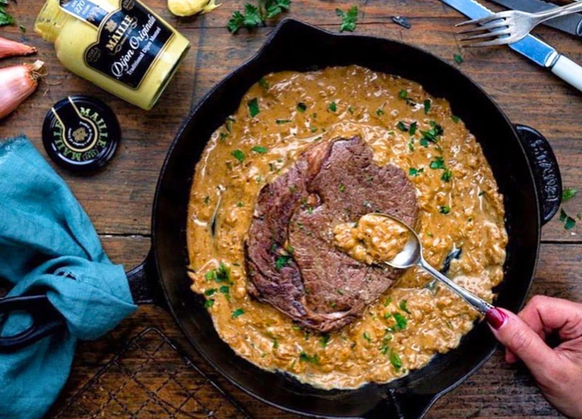 A hand holds a spoon over a skillet with steak in Diane sauce, garnished with herbs.