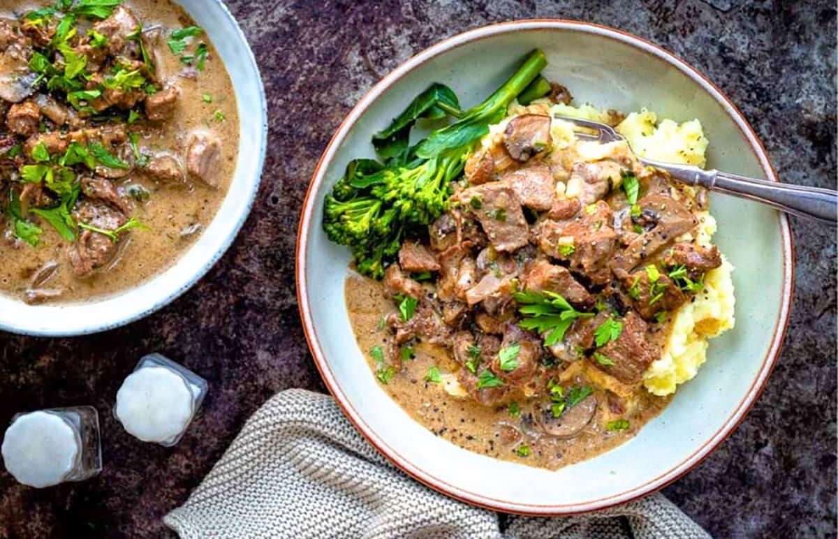 A bowl of creamy steak diane with mushrooms served over mashed potatoes, garnished with parsley and accompanied by steamed broccoli. A fork and napkin are nearby beside a second dish of the same meal.