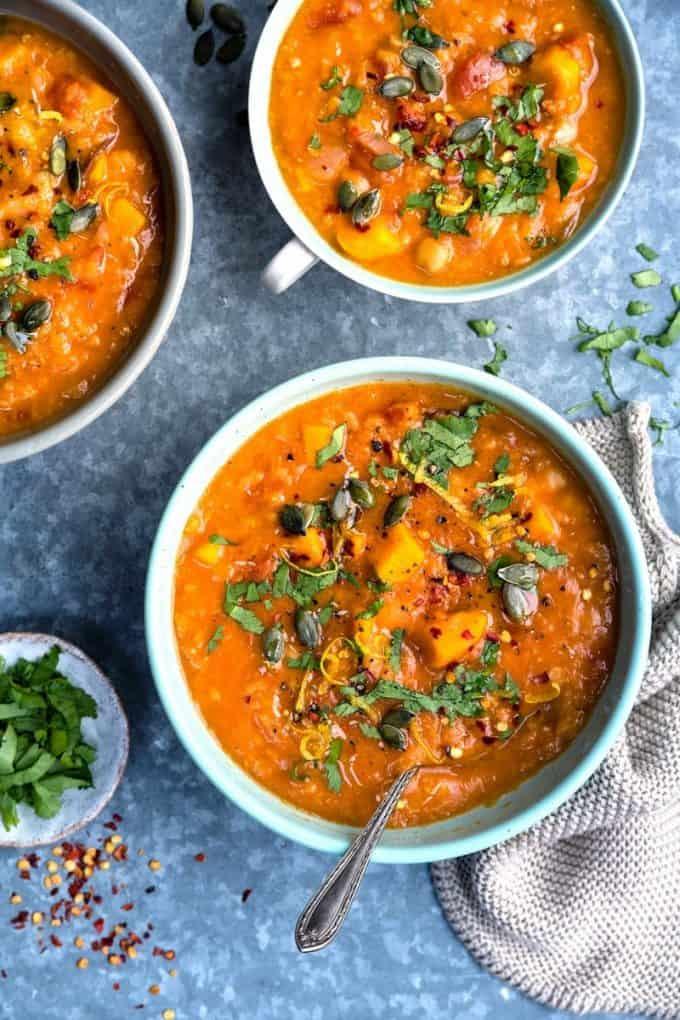 Three bowls of hearty sweet potato chickpea red lentil soup, garnished with chopped fresh herbs and pumpkin seeds, sit on a blue surface with a spoon in one bowl and a small dish of chopped herbs nearby.