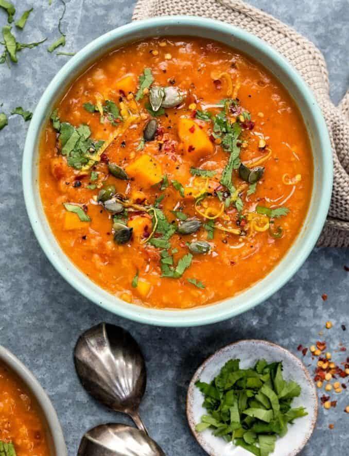 A bowl of sweet potato chickpea red lentil soup, orange-hued and garnished with fresh herbs, pumpkin seeds, and spices, sits beside a small dish of chopped herbs and two metal spoons on a gray surface.