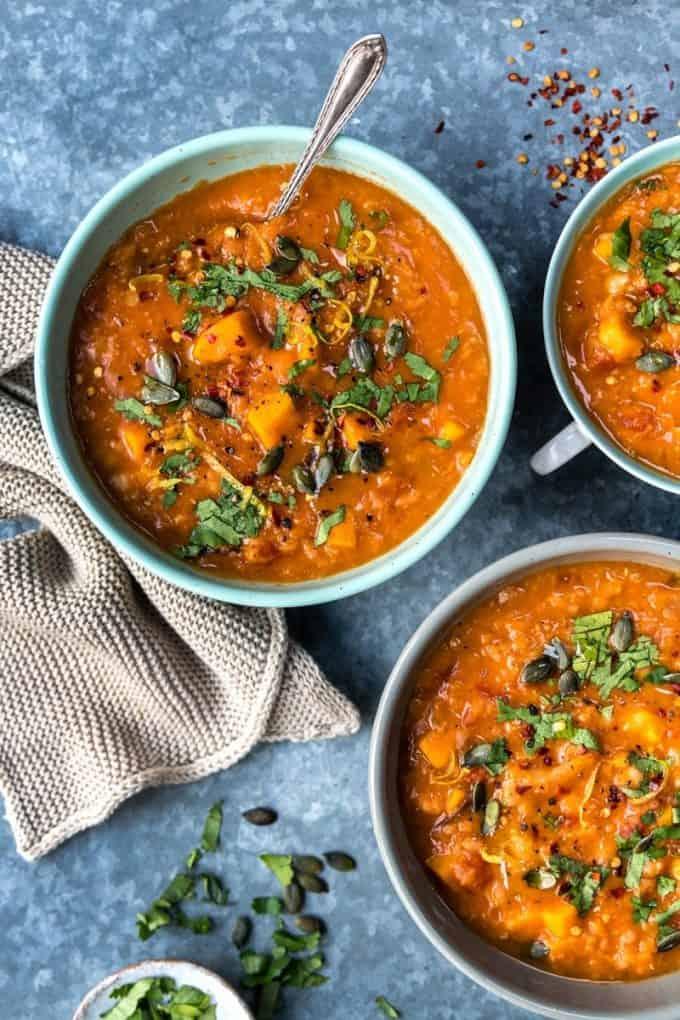 Three bowls of hearty sweet potato chickpea red lentil soup, garnished with fresh herbs and seeds, sit on a blue surface. A spoon rests in one bowl, while a beige knitted cloth and scattered herbs and seeds complete the cozy scene.