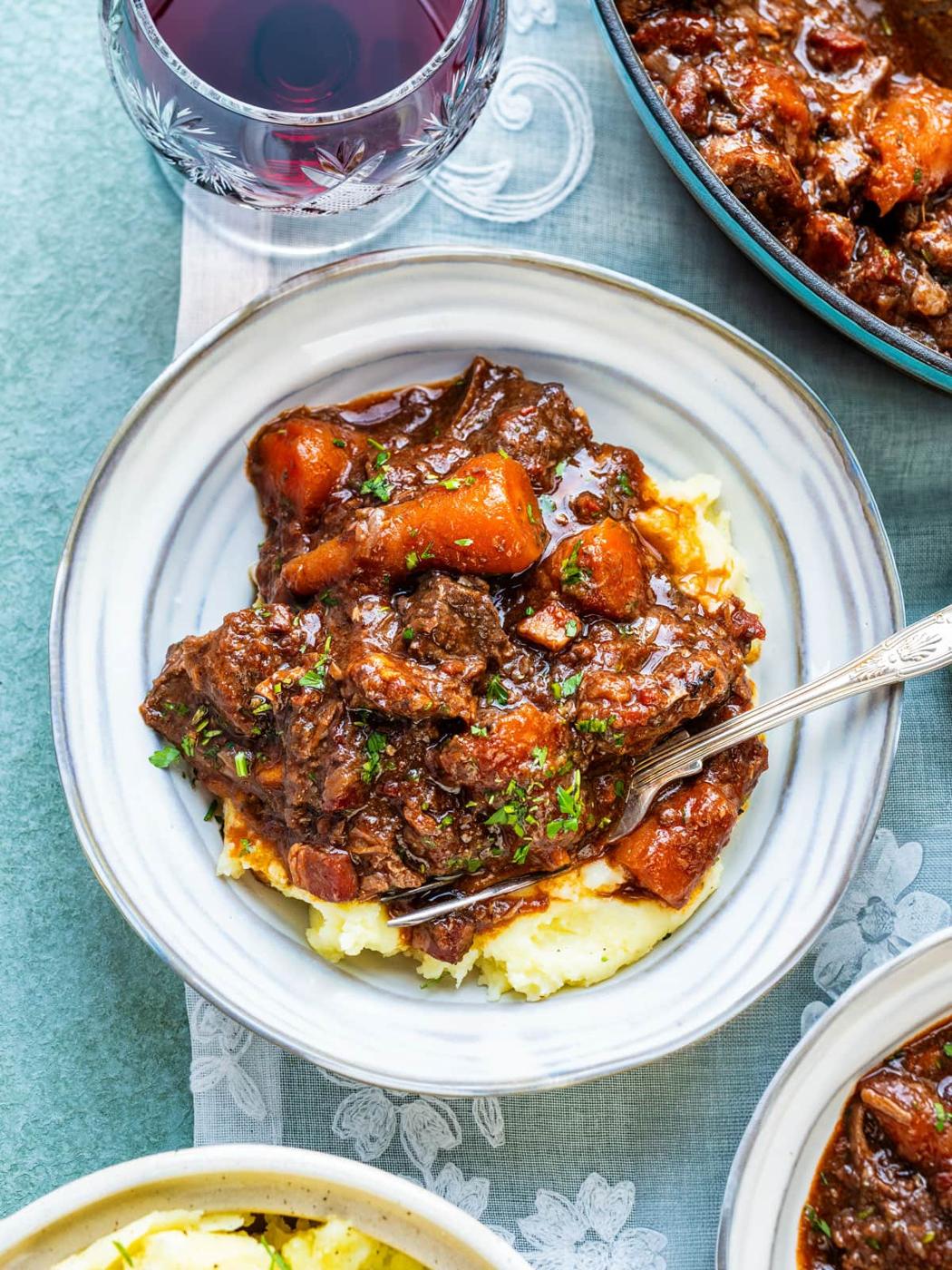 A plate of Provencal beef stew with carrots and rich brown sauce served over mashed potatoes, garnished with chopped herbs, sits next to a glass of red wine and a bowl of food on a blue tablecloth.
