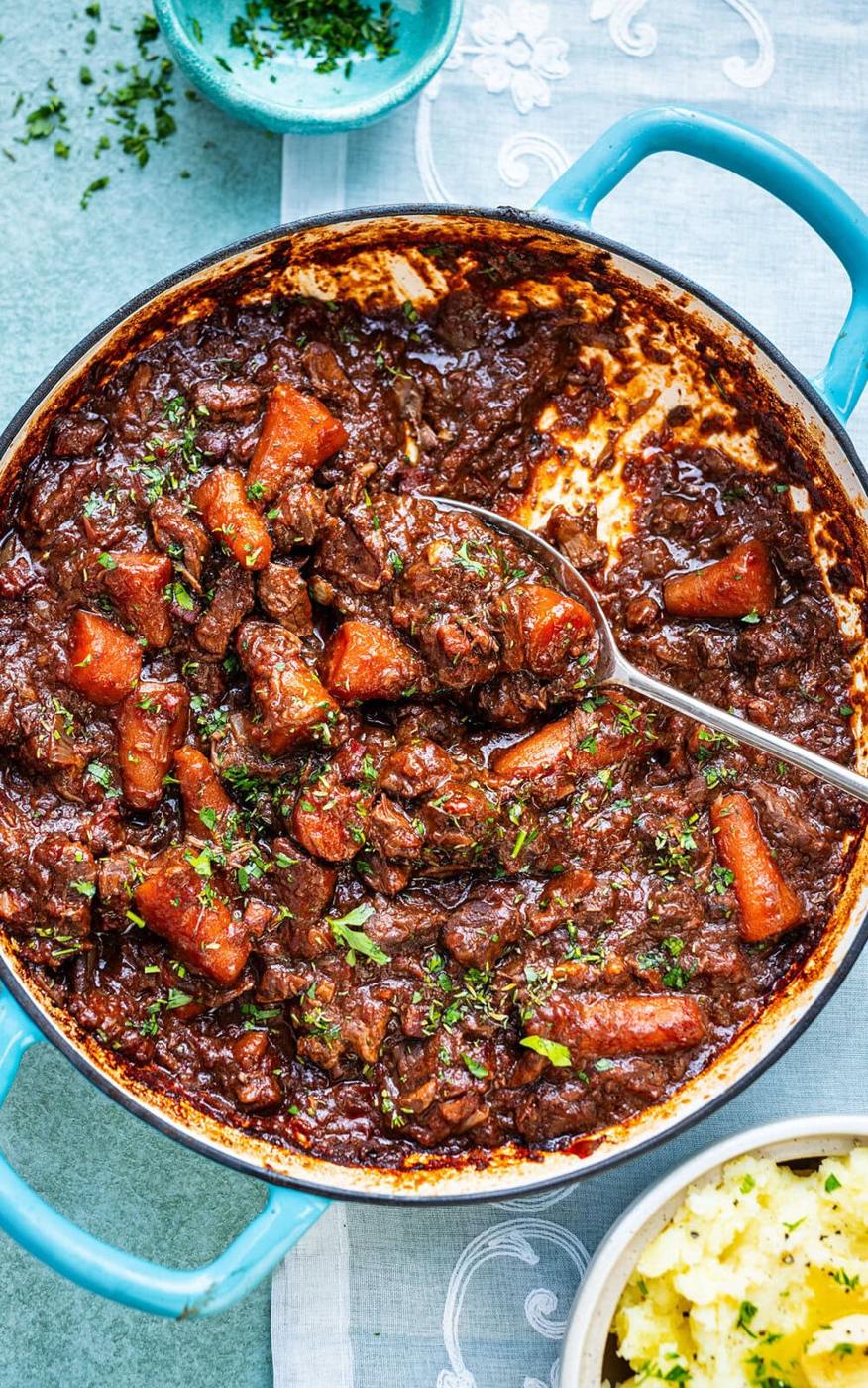 A blue pot filled with Provencal beef stew featuring chunks of beef, carrots, and a rich, dark sauce topped with chopped herbs. A serving spoon rests inside the pot, and mashed potatoes are visible beside it.