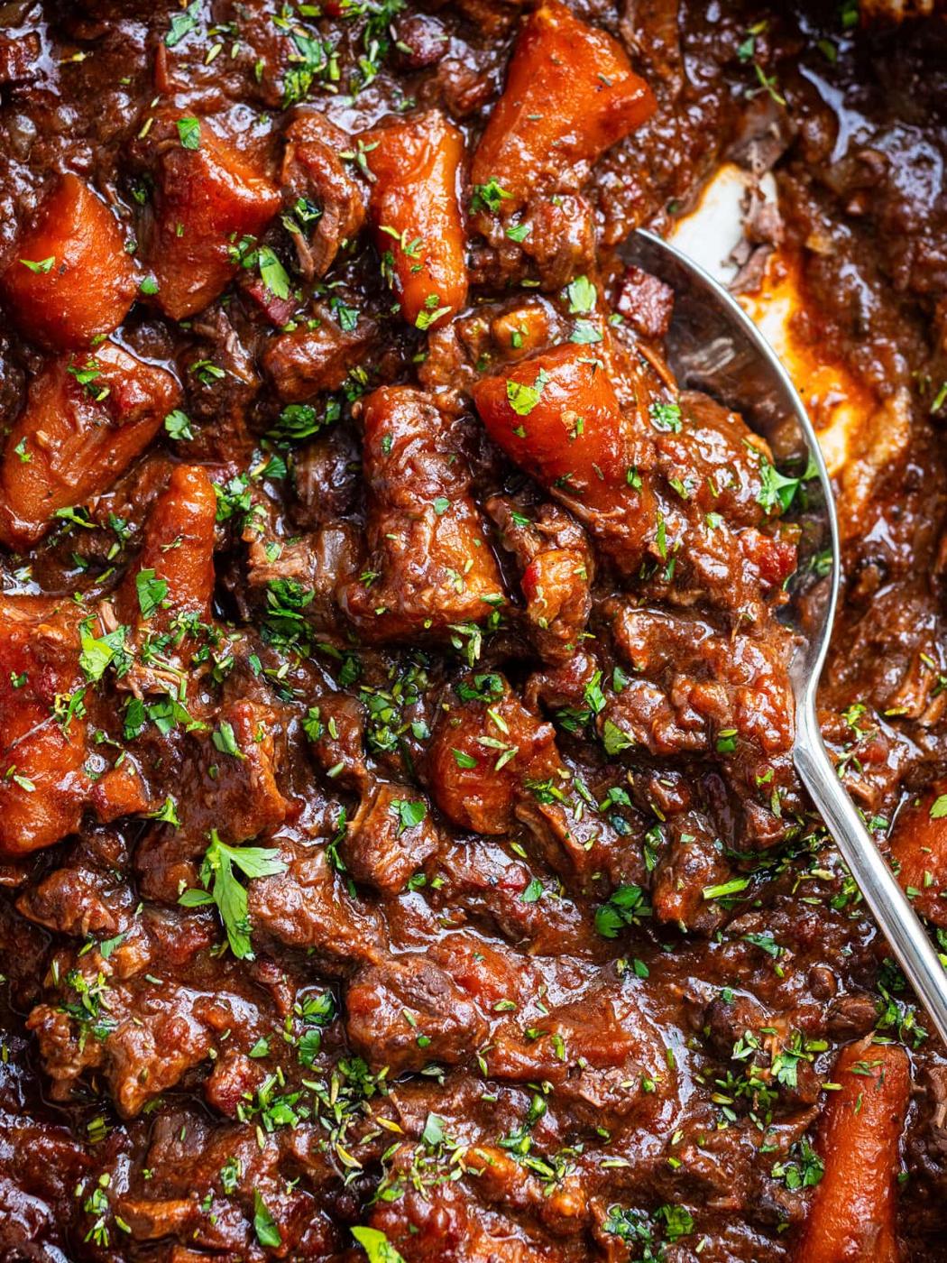 Close-up of a hearty beef stew with chunks of beef, carrots, and a rich brown sauce, garnished with chopped parsley. A metal spoon is partially submerged in the thick, flavorful mixture.