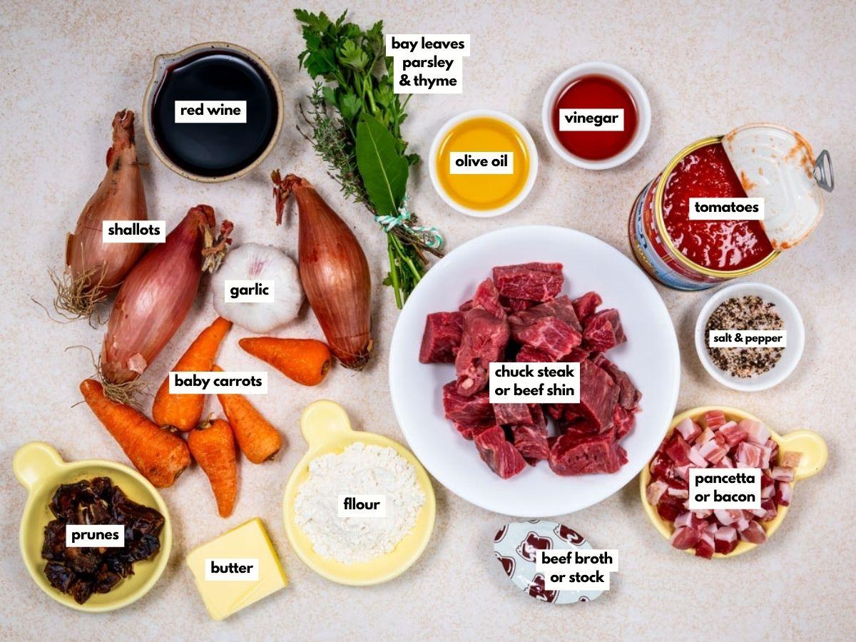 A flat lay of Provençal beef Daube stew with celeriac mash ingredients, including cubed chuck steak, baby carrots, shallots, garlic, prunes, pancetta, red wine, tomato, herbs, and spices.