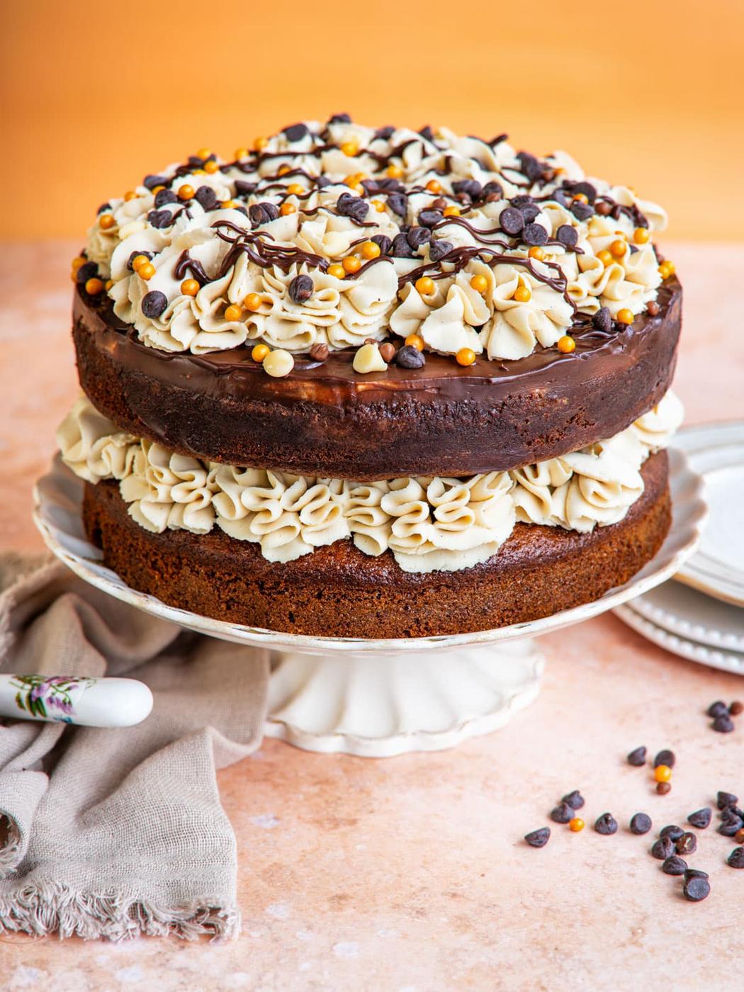 A two-layer Pumpkin Chocolate Chip Cake with cream filling and decorative cream swirls on top, garnished with chocolate chips, gold sprinkles, and a drizzle of chocolate, displayed on a cake stand.
