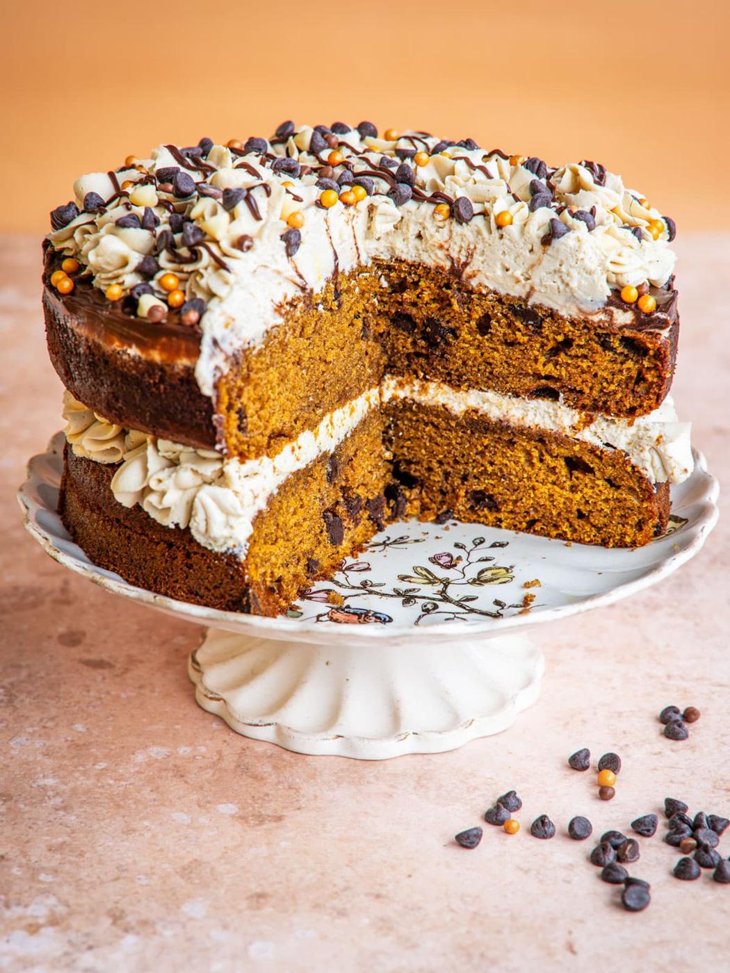 A two-layer pumpkin cake with cream frosting, chocolate chips, and sprinkles sits on a decorative cake stand. A large slice has been cut, revealing the moist, dense interior with chocolate chips.