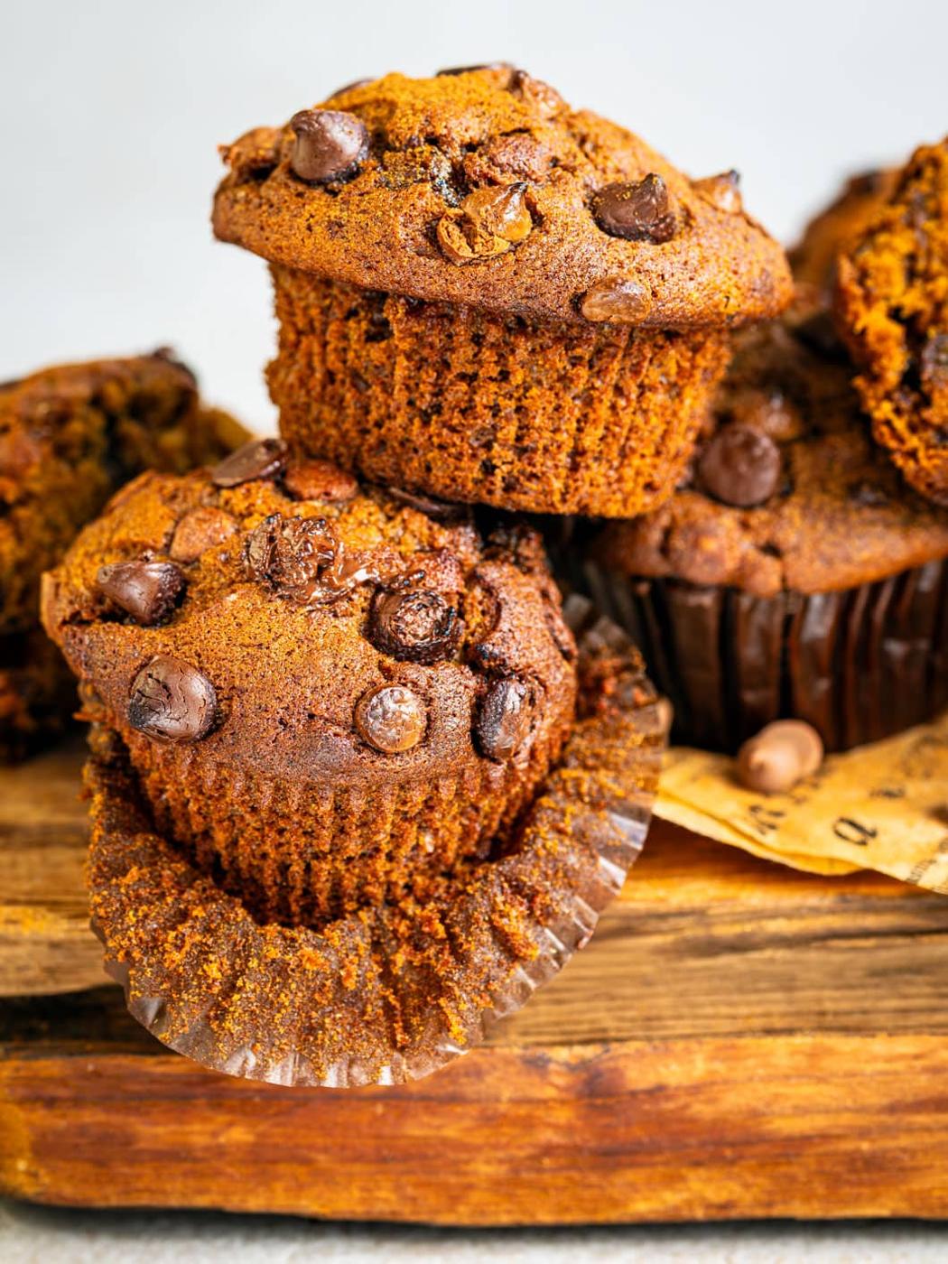 Two pumpkin chocolate chip muffins are stacked on a rustic wooden surface, with the bottom muffin partially unwrapped, revealing its moist texture. More muffins are visible in the background.