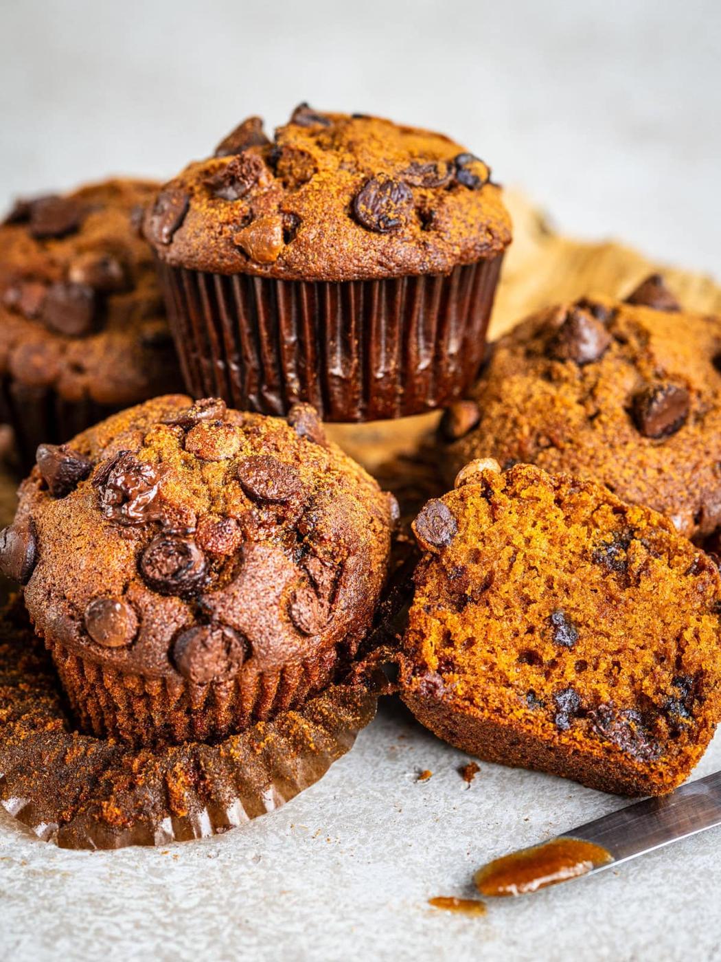Chocolate chip pumpkin muffins are arranged on a light surface; one muffin is cut open to show a moist, chocolatey interior. Chocolate chips are visible on top and inside the muffins, with crumbs scattered nearby.