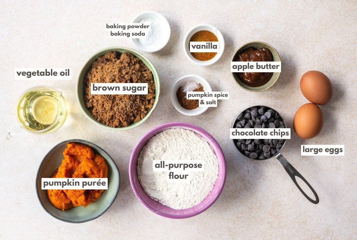 Top-down view of baking ingredients for a pumpkin chocolate chip muffin recipe: vegetable oil, brown sugar, pumpkin purée, all-purpose flour, baking powder, baking soda, pumpkin spice, salt, vanilla, apple butter, chocolate chips, and two large eggs.