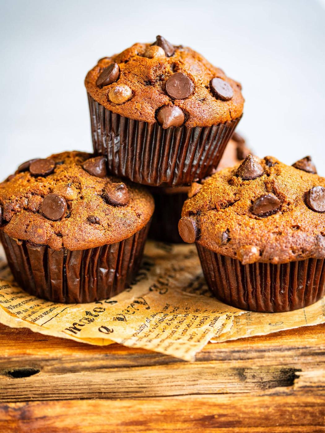 Three chocolate chip muffins in brown wrappers are stacked on vintage newspaper on a rustic wooden surface, with golden tops and scattered chocolate chips visible.