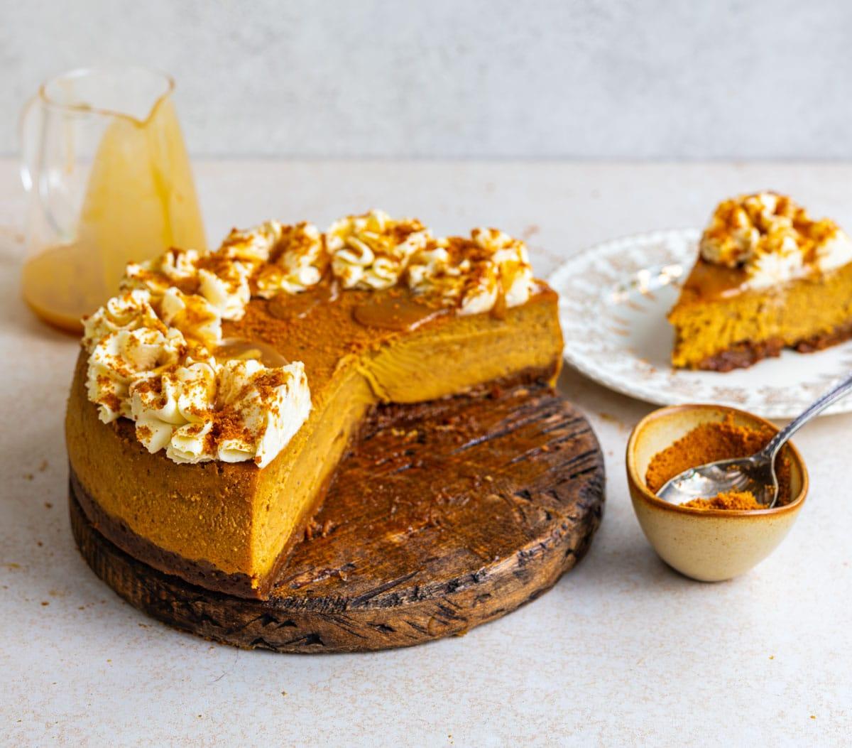 Pumpkin pie cheesecake with a Biscoff crust on a wooden board, a slice cut away to show the creamy filling, with a slice on a plate, a bowl of Biscoff crumbs and a jug of caramel sauce.