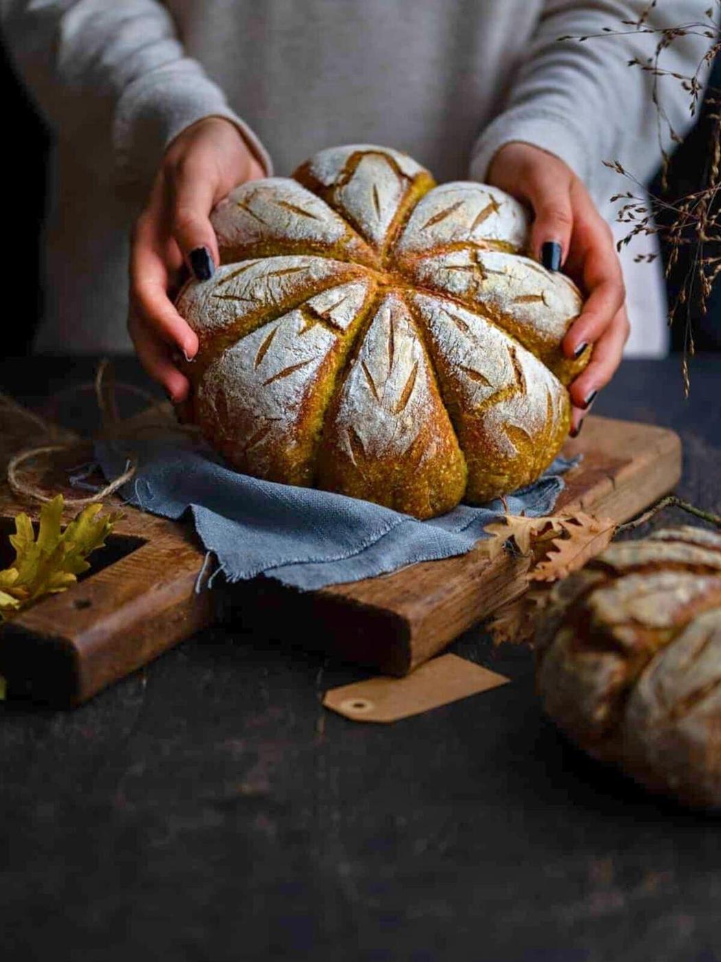 Hands lifting a baked pumpkin sourdough loaf, scored into eight segments, above a wooden board and blue linen cloth.