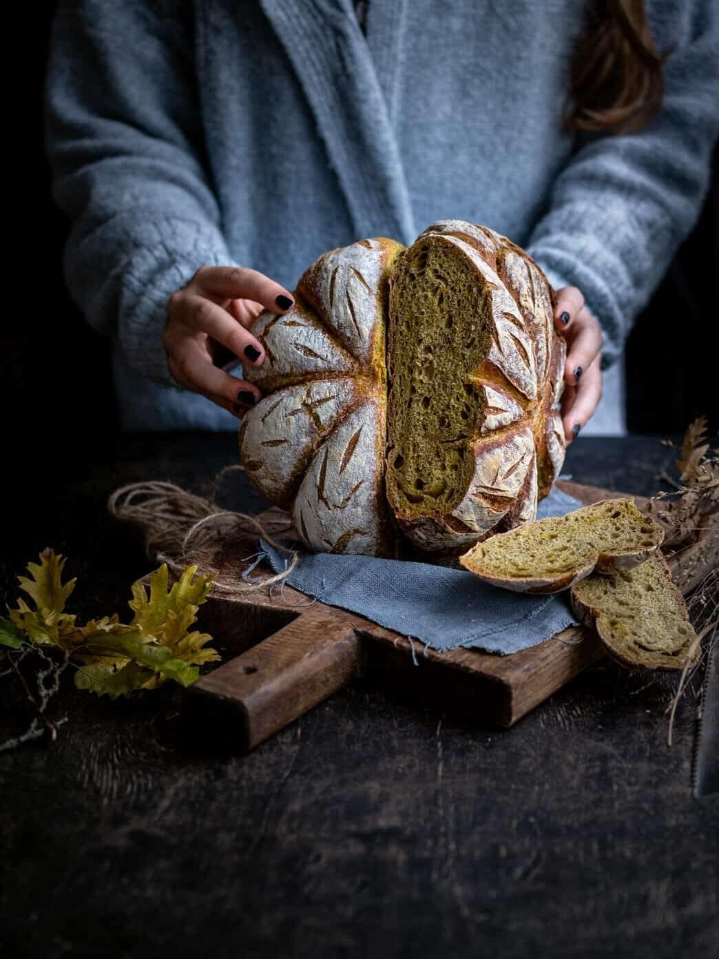 A wedge being lifted from a pumpkin-shaped sourdough loaf to show the soft golden crumb inside, with slices on a wooden board.