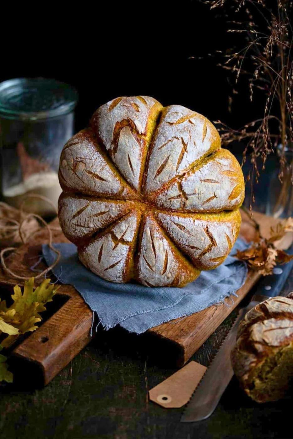 A baked pumpkin sourdough loaf, scored into eight segments, standing on a blue cloth over a wooden board against a dark background.