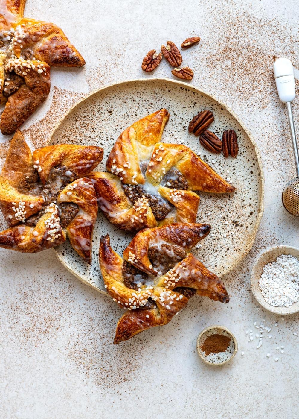 Pinwheel-shaped Danish pastries with pearl sugar sit on a beige plate, surrounded by pecans, a small bowl of flour, cinnamon, and a pastry tool on a light textured surface.