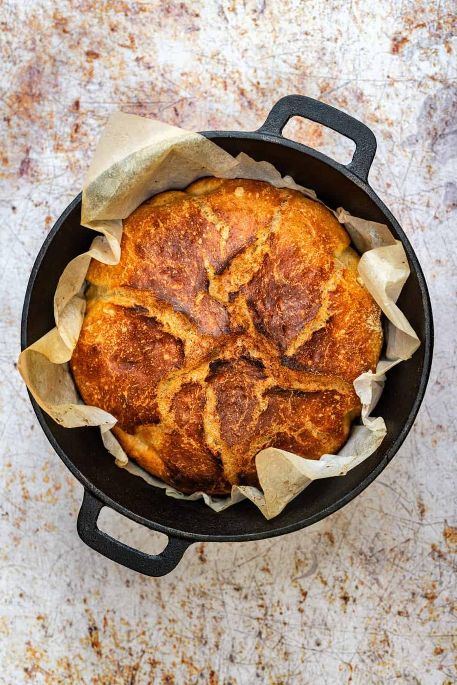 A round loaf of quick sourdough bread with yeast, golden-brown with a rustic crust, sits on parchment paper in a black cast iron pot, viewed from above on a textured surface.