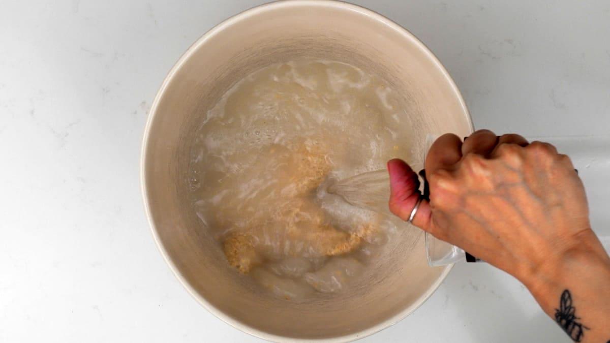 A hand pours water from a clear pitcher into a beige bowl containing sourdough starter, viewed from above on a white surface.