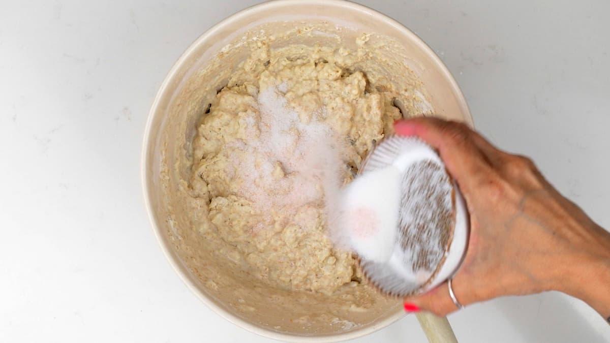 A hand pours granulated sugar and salt from a small bowl into a larger bowl containing thick, beige sourdough batter. The bowl rests on a white surface.