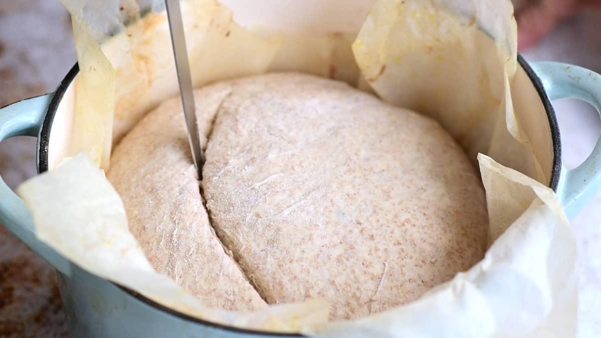 A close-up of sourdough bread dough in a parchment-lined pot, being scored with a knife before baking.